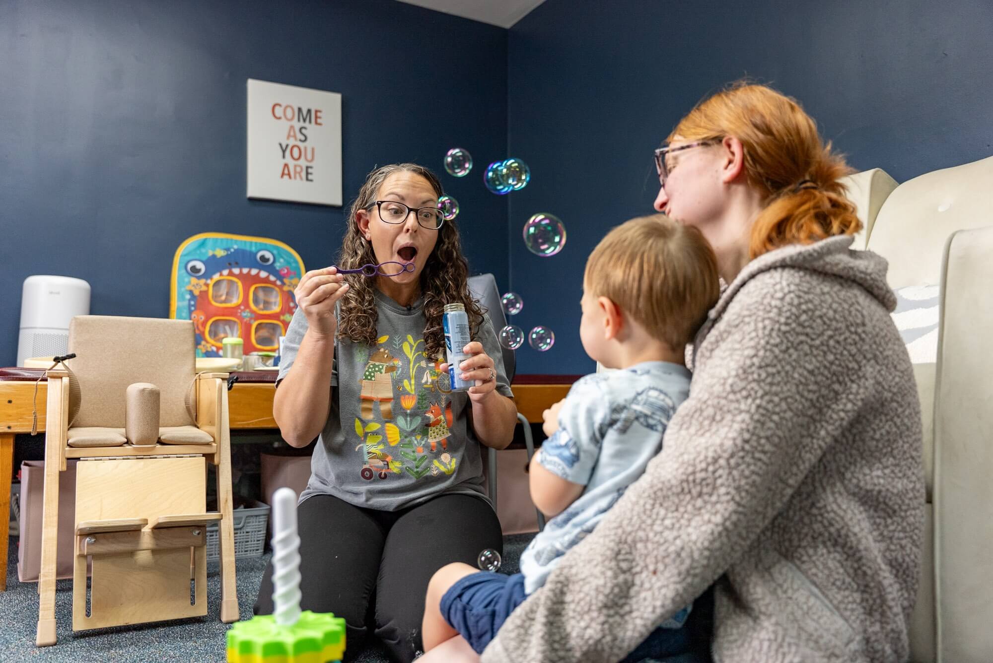 Therapist blowing bubbles for a toddler sitting on a parent’s lap