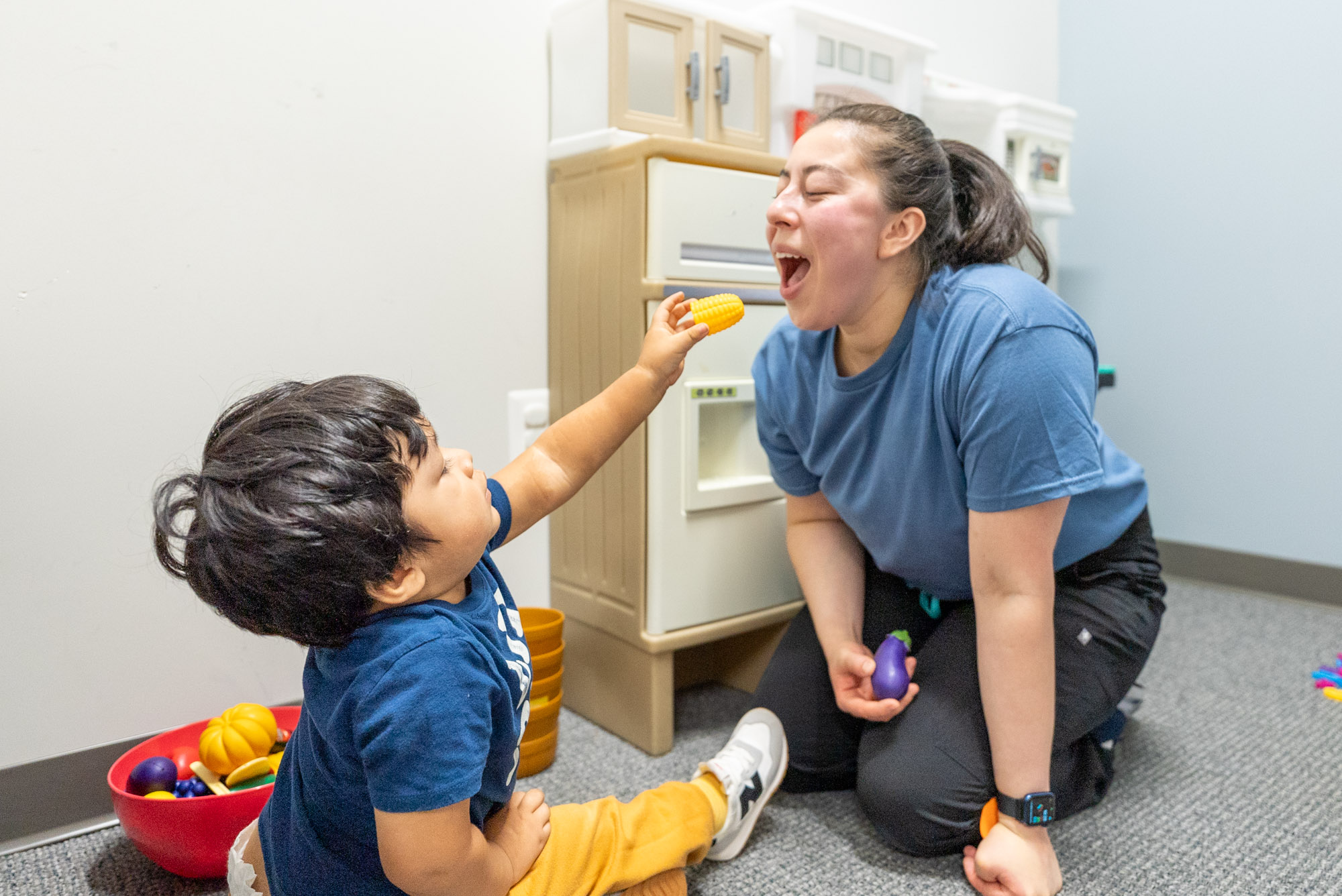Child offering a toy to a smiling therapist in a play kitchen