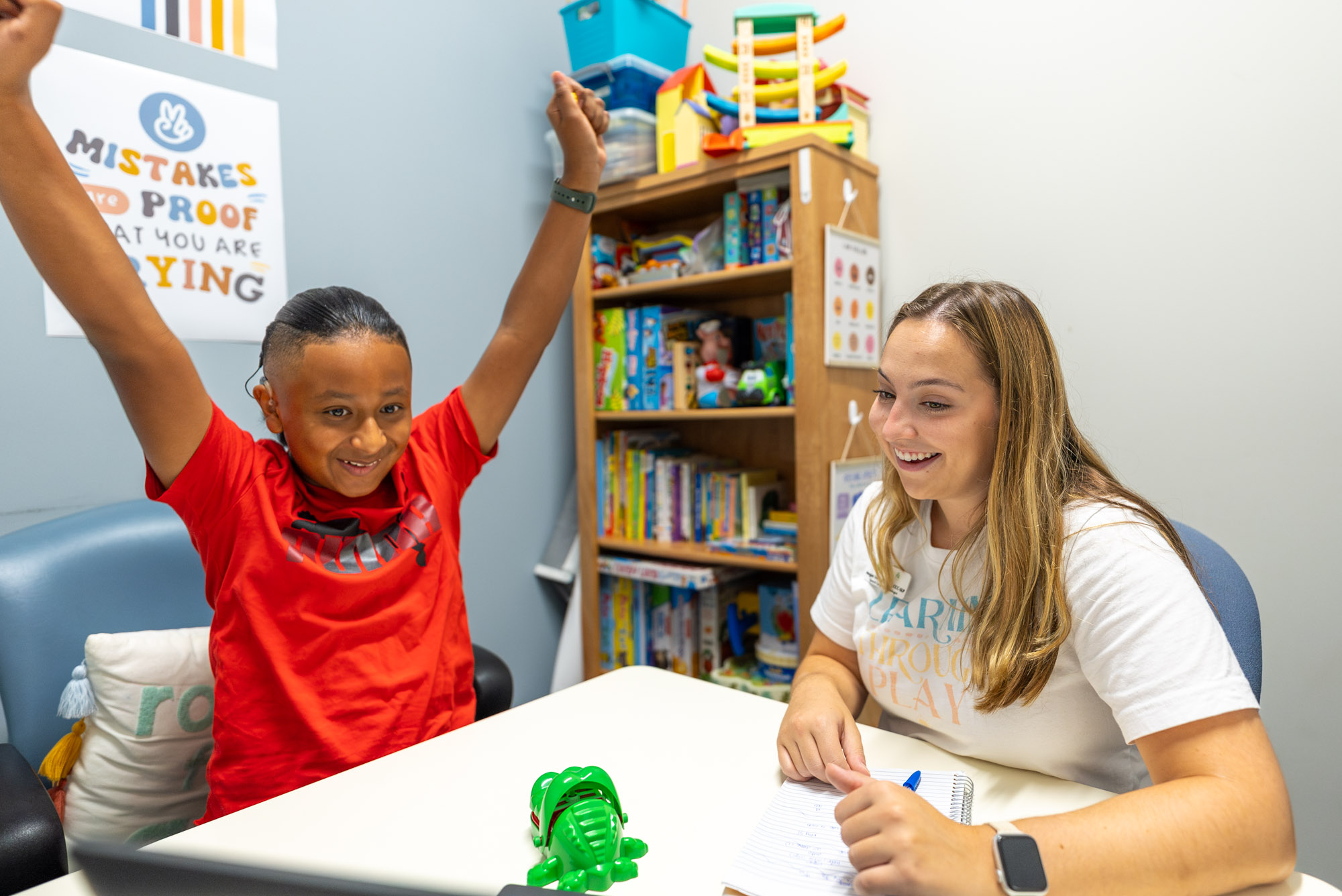 Child celebrating with arms raised at a table beside a smiling therapist